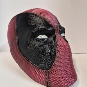 May include: A full-head mask featuring a textured, deep red surface with prominent black eye patches. The eye areas are covered with a white, fine-mesh material. The overall surface has a consistent, small-scale grid pattern throughout.