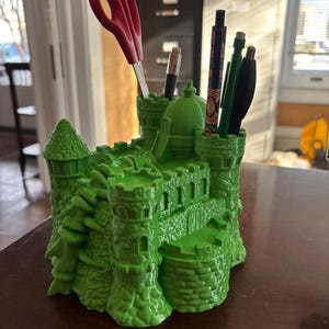 May include: A bright green castle-shaped desk organizer with multiple compartments. Red scissors, pens, and pencils are stored inside. The organizer has turrets, walls, and a detailed architectural design, perfect for a desk or office.