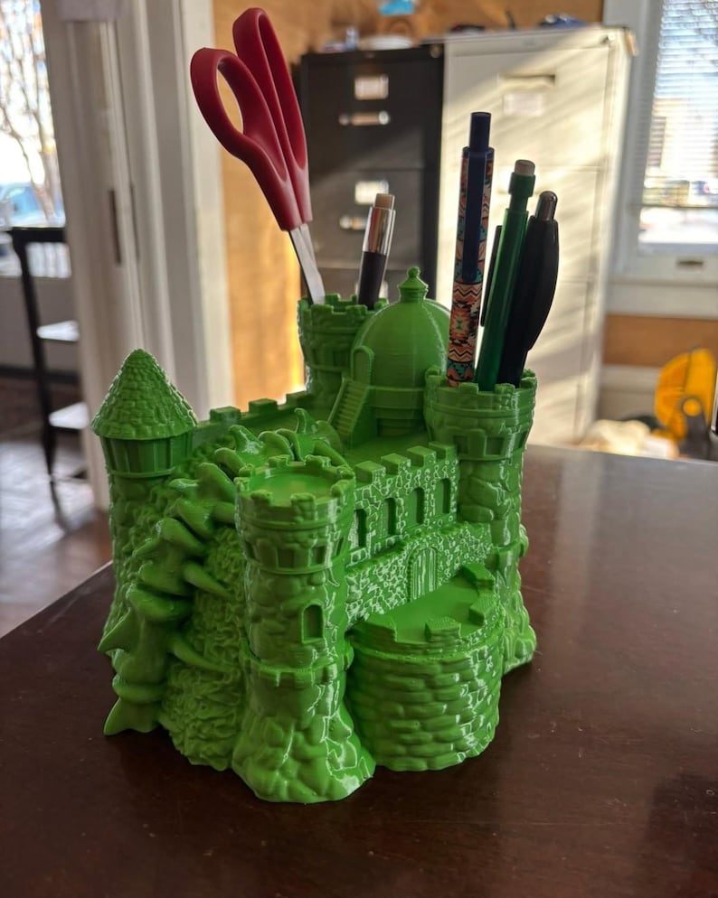 May include: A bright green castle-shaped desk organizer with multiple compartments. Red scissors, pens, and pencils are stored inside. The organizer has turrets, walls, and a detailed architectural design, perfect for a desk or office.