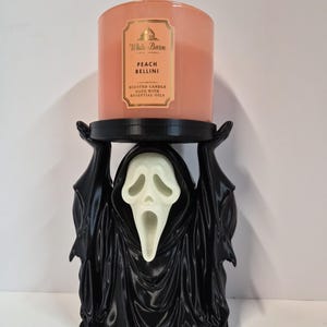 Könnte beinhalten: Eine pfirsichfarbene White Barn Peach Bellini Duftkerze im Glas steht auf einem schwarzen Kerzenhalter in Form einer gehüllten Figur mit einer weißen, länglichen, geisterhaften Maske. Die Figur hält die Arme hoch, um den Kerzensockel zu stützen.