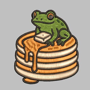May include: An embroidered patch featuring a green frog sitting atop a stack of pancakes. The pancakes are golden brown with syrup dripping down the side. A square of butter sits on top of the pancakes.
