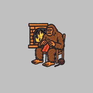 Op de afbeelding: Geborduurde patch met een bruine Bigfoot die voor een open haard zit te breien met rood garen. De Bigfoot heeft een serieuze uitdrukking. Een bol wol en een breinaald liggen op de grond.