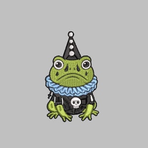 May include: Embroidered patch featuring a green frog wearing a black outfit with a skull, a blue ruffled collar, and a black hat with white dots. The frog has large, expressive eyes and a sad expression.