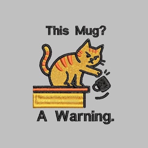 May include: Embroidered design on a gray background. The image features a yellow cat with orange stripes knocking over a black mug. Text above reads "This Mug?" and below "A Warning."