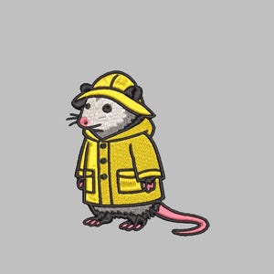 Puede incluir: Diseño bordado de una zarigüeya con impermeable amarillo y sombrero a juego. La zarigüeya tiene pelaje gris, nariz rosa y cola rosa. El impermeable tiene botones y bolsillos negros. El diseño está sobre un fondo gris claro.