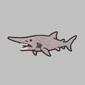 May include: An embroidered patch featuring a light pink goblin shark with a long snout, sharp white teeth, and black outlines. The shark is depicted in a side profile view with visible gills, fins, and a tail, rendered in detailed thread work.