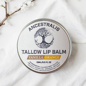 Puede incluir: Una lata plateada de bálsamo labial Ancestralis de sebo con sabor a vainilla y naranja. La lata presenta un árbol con raíces y un círculo a su alrededor. Los ingredientes se enumeran en la parte inferior de la lata: "Ingredientes: Sebo de res alimentado con pasto, aceite de jojoba orgánico, cera de abejas, vainilla orgánica y aceites esenciales de naranja".