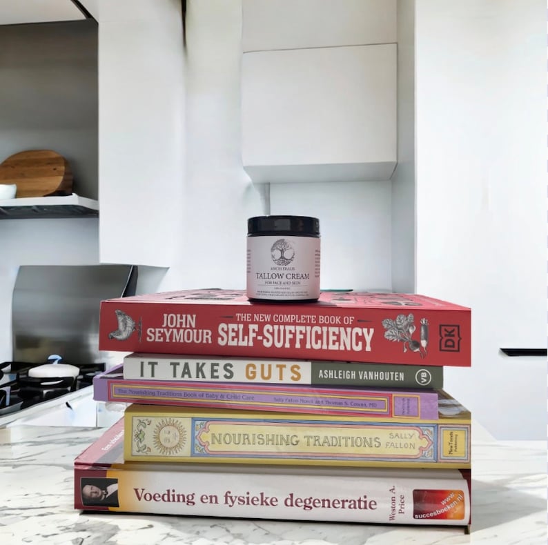 Peut inclure: Une pile de quatre livres avec un petit pot de cr&egrave;me de suif sur le dessus. Le livre du haut est rouge et porte le titre "The New Complete Book of Self-Sufficiency" de John Seymour. Le deuxi&egrave;me livre est bleu et porte le titre "It Takes Guts" d'Ashleigh VanHouten. Le troisi&egrave;me livre est jaune et porte le titre "Nourishing Traditions" de Sally Fallon. Le livre du bas est rouge et porte le titre "Voeding en fysieke degeneratie" de Weston A. Price. Le pot de cr&egrave;me de suif est blanc avec une &eacute;tiquette noire qui dit "Ancestral Tallow Cream for Hair and Skin".