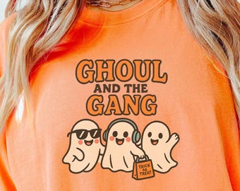 May include: Bright orange t-shirt featuring the words "GHOUL and the GANG" above a graphic of three cartoon ghosts. One ghost wears sunglasses, another has headphones, and the third carries a "Trick or Treat" bag. A Halloween-themed garment.