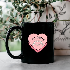 May include: Black coffee mug with a pink heart design that says 'til death' with skeleton hands forming a heart shape inside the heart.