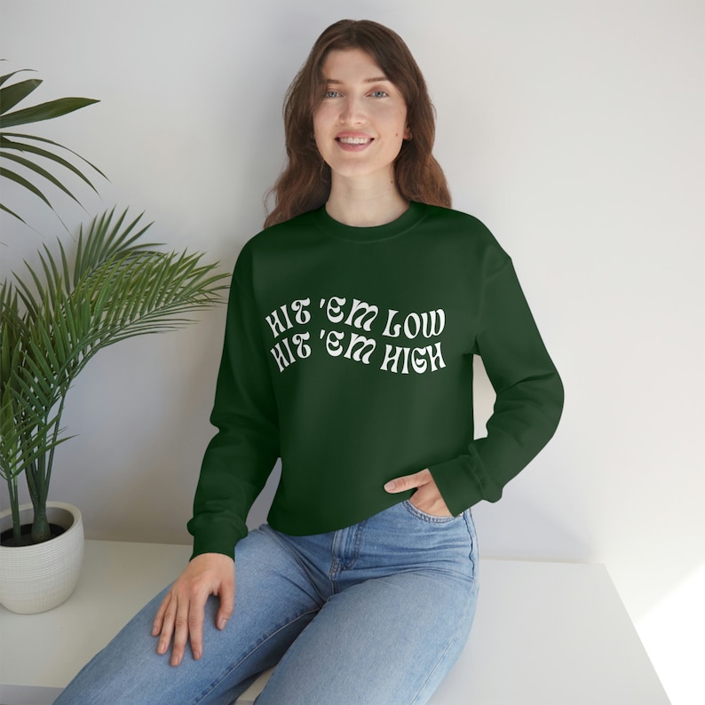 Pu&ograve; includere: Una felpa verde scuro con testo bianco che recita "Hit 'em Low Hit 'em High".