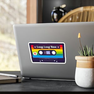 May include: A sticker of a retro cassette tape with a rainbow stripe design. The text "Long, Long Time" is displayed across the top in white, with a red heart on each side. The letter "A" is in a red square.