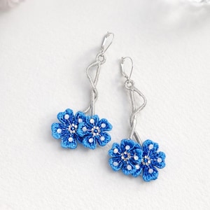 May include: A pair of dangle earrings featuring bright blue flower designs with white accents. The floral elements are connected to silver-toned, twisted wire stems. The earrings are set against a white background.