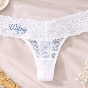 Op de afbeelding: Witte kanten string met het woord "Wifey" in lichtblauw geborduurd. De delicate lingerie heeft een bloemen kanten patroon. Een romantisch en vrouwelijk stuk.
