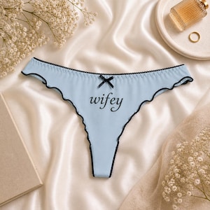 Op de afbeelding: Lichtblauwe string met zwarte rand en een zwarte strik. Het woord "wifey" staat in cursief op de voorkant. Het ondergoed heeft een geschulpte rand.