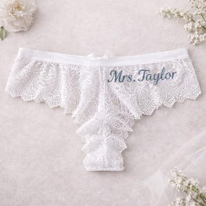 Puede incluir: Ropa interior de encaje blanco con el texto bordado "Mrs. Taylor". La delicada lencería presenta un borde festoneado y un pequeño lazo en la cintura. El fondo incluye flores blancas.