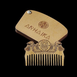 May include: A wooden comb with an intricate design featuring a Viking warrior's face and two birds. The comb is made of light brown wood and has a natural finish. The comb is packaged in a wooden box with the word "ARHAIKA" engraved on it.