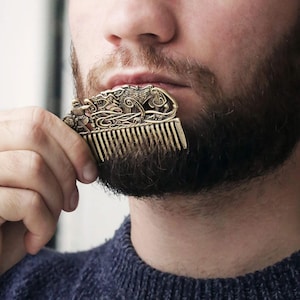 May include: A gold-toned comb with an intricate design featuring a wolf and knotwork. The comb is being held by a person's hand.