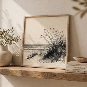 Könnte beinhalten: Ein gerahmter Schwarz-Weiß-Druck einer Strandszene. Das Kunstwerk zeigt eine Küstenlandschaft mit Sanddünen, hohem Gras und dem Meer. Der Druck befindet sich in einem hellen Holzrahmen und steht auf einem Holzregal mit Büchern und einer Vase.