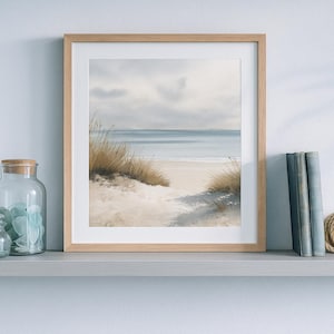 Op de afbeelding: Een ingelijste aquarelprint van een strandscène met zandduinen en de oceaan. Het kunstwerk zit in een licht houten lijst. Decoratieve glazen potten en boeken staan op een plank.