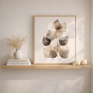 Op de afbeelding: Een ingelijste kunstdruk met een abstract aquarelontwerp van gelaagde bladeren in bruine en beige tinten. Het kunstwerk wordt tentoongesteld op een houten plank met een vaas, boeken en schelpen.