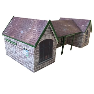 以下が含まれることがあります： 石造りのファサードと緑色の屋根を持つ、厚紙製の模型の駅キット。駅には「Carlisle Train Station West」と書かれた看板があります。