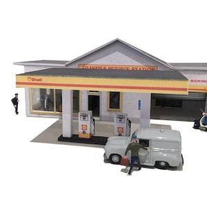 Puede incluir: Un modelo de una gasolinera vintage con una furgoneta blanca que se acerca a las bombas de gasolina. La gasolinera tiene un letrero que dice "Frankie's Service Station" y un logotipo de Shell. Hay una scooter estacionada frente a la puerta del garaje.