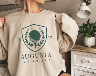 May include: A beige sweatshirt with a green graphic of a golf ball on a tee inside a shield with a laurel wreath. The text "19 32 AUGUSTA GEORGIA" is printed below the graphic.