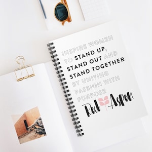 May include: A white spiral bound notebook with the text "Inspire Women to Stand Up, Stand Out and Stand Together by Uniting Passion with Purpose" and the logo "Red Aspen" with a pink flower.
