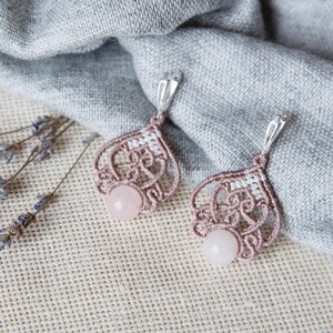 May include: A pair of handmade macramé earrings with a pink rose quartz bead. The earrings are a light mauve colour with a silver-coloured hook. Dried lavender sprigs and a grey linen cloth are in the background.