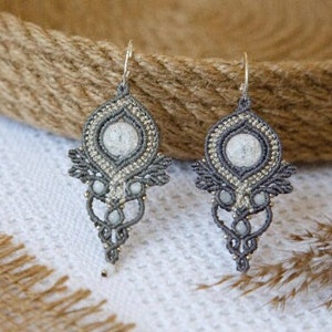 May include: Two grey macrame earrings, each featuring a central white stone and gold-coloured detailing. The earrings have an ornate, teardrop design. They are presented on a white surface, with a woven basket visible in the background.