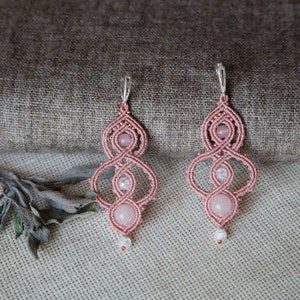 May include: Handmade pink macrame earrings with rose quartz and clear crystal details. The earrings have a woven pattern, a central rose quartz bead, and smaller crystal beads. Silver-tone ear wires complete the design.