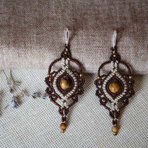 May include: A pair of handcrafted macrame earrings. The earrings feature a brown and cream-colored woven design with a central, round, speckled stone. Small gold-colored beads accent the design. The earrings hang from silver-colored hooks.