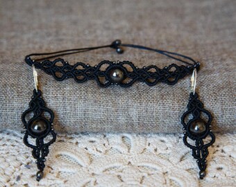 May include: A black macrame jewelry set featuring a choker necklace and matching earrings. Each piece has a central dark gray bead and intricate knotwork. The earrings have silver-colored hooks.
