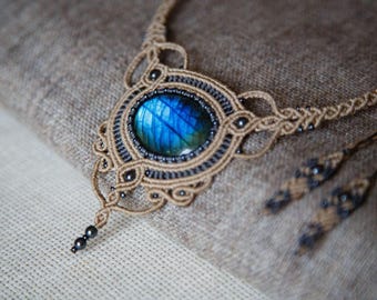 May include: A handcrafted macrame necklace featuring a large, oval, blue gemstone. The intricate design incorporates black beads and a tan cord, creating a bohemian aesthetic. The necklace is displayed on a textured, neutral-toned fabric.