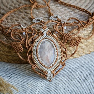 May include: A handcrafted macrame necklace featuring a central oval stone, surrounded by intricate woven patterns in shades of brown and cream. The necklace has a bib-style design with delicate details and small white beads.