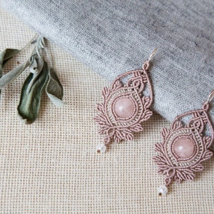 Puede incluir: Un par de pendientes de macramé hechos a mano con una piedra central de cuarzo rosa claro. Los pendientes tienen un diseño calado detallado en color rosa empolvado, con una pequeña cuenta blanca en la parte inferior. Se exhiben sobre una superficie de lino neutro.