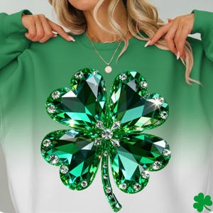 May include: Green sweatshirt featuring a large, sparkling shamrock design. The shamrock is a vibrant green with crystal accents, set against a gradient background that fades from green to white. A small shamrock graphic is in the lower right corner.