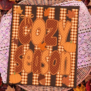 May include: A rectangular sign featuring a brown and orange gingham background with vertical stripes. The text COZY SEASON is written in large, polka-dotted letters. The design is decorated with orange pumpkins, an acorn, and autumn leaves.