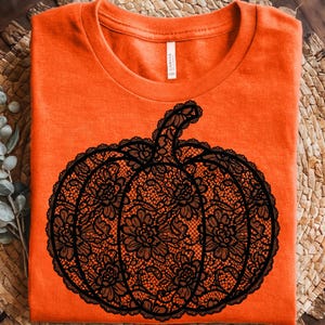 May include: A bright orange t-shirt featuring a large, central graphic of a pumpkin. The pumpkin design is rendered in a detailed black lace pattern, showcasing intricate floral motifs and scalloped edges throughout the entire shape.