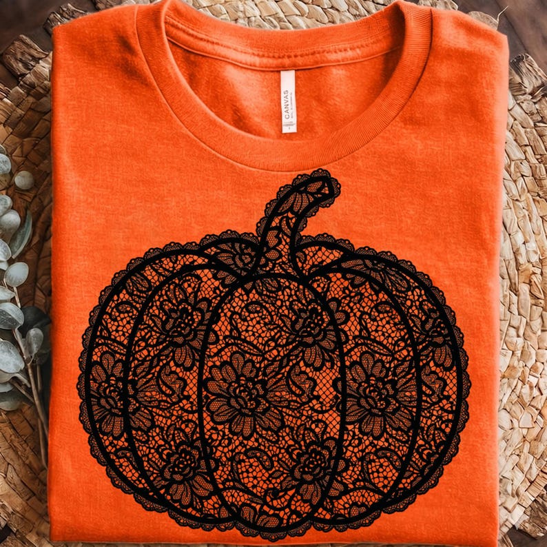 May include: A bright orange t-shirt featuring a large, central graphic of a pumpkin. The pumpkin design is rendered in a detailed black lace pattern, showcasing intricate floral motifs and scalloped edges throughout the entire shape.