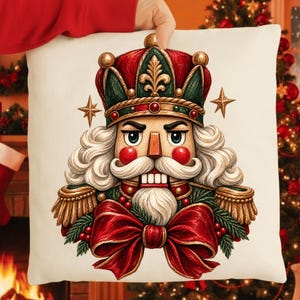 May include: A square white throw pillow featuring a detailed illustration of a nutcracker wearing a green and red crown with gold accents. The nutcracker has white hair, a mustache, a beard, and a large red bow with green pine needles.