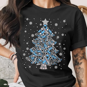 May include: Black t-shirt featuring a Christmas tree design. The tree is composed of various blue and gray symbols, including hands, ribbons, and gender symbols, with a gray star on top. White snowflakes are scattered around the tree.
