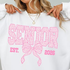 Puede incluir: Sudadera blanca con la palabra "SENIOR" en letras rosas grandes con estampado de leopardo. Debajo, un lazo rosa. El texto "EST. 2026" tambi&eacute;n es rosa.