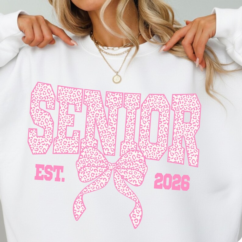 Puede incluir: Sudadera blanca con la palabra "SENIOR" en letras rosas grandes con estampado de leopardo. Debajo, un lazo rosa. El texto "EST. 2026" tambi&eacute;n es rosa.