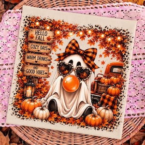 Peut inclure: Une plaque décorative carrée représentant un fantôme portant un nœud à carreaux et des lunettes en forme de cœur, soufflant une bulle orange. Le décor inclut des citrouilles, une lanterne, un camion orange et un panneau en bois avec le texte HELLO FALL COZY DAYS WARM DRINKS AND GOOD VIBES entouré de feuilles d'automne.