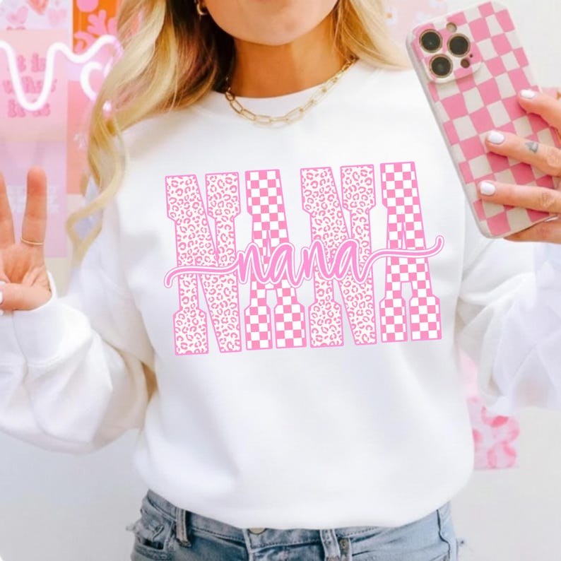 May include: White sweatshirt with the word "NANA" in pink, featuring a mix of leopard print and checkered patterns. The word "nana" is written in cursive below the word "NANA". A pink and white checkered phone case is also visible.
