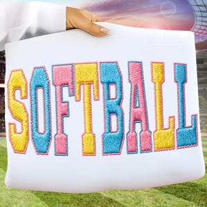 May include: A white pillow with the word "SOFTBALL" embroidered in colorful thread. The letters are in shades of yellow, pink, and blue. The pillow is held up by a person in a white shirt, with a green field and stadium in the background.
