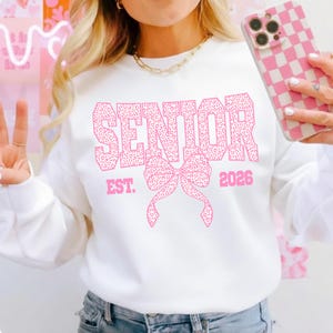 Puede incluir: Sudadera blanca con la palabra "SENIOR" en letras rosas con estampado de leopardo, un lazo rosa y el a&ntilde;o "2026". Una sudadera de estilo casual, ideal para una celebraci&oacute;n de graduaci&oacute;n.
