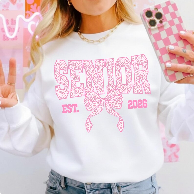 Puede incluir: Sudadera blanca con la palabra "SENIOR" en letras rosas con estampado de leopardo, un lazo rosa y el a&ntilde;o "2026". Una sudadera de estilo casual, ideal para una celebraci&oacute;n de graduaci&oacute;n.